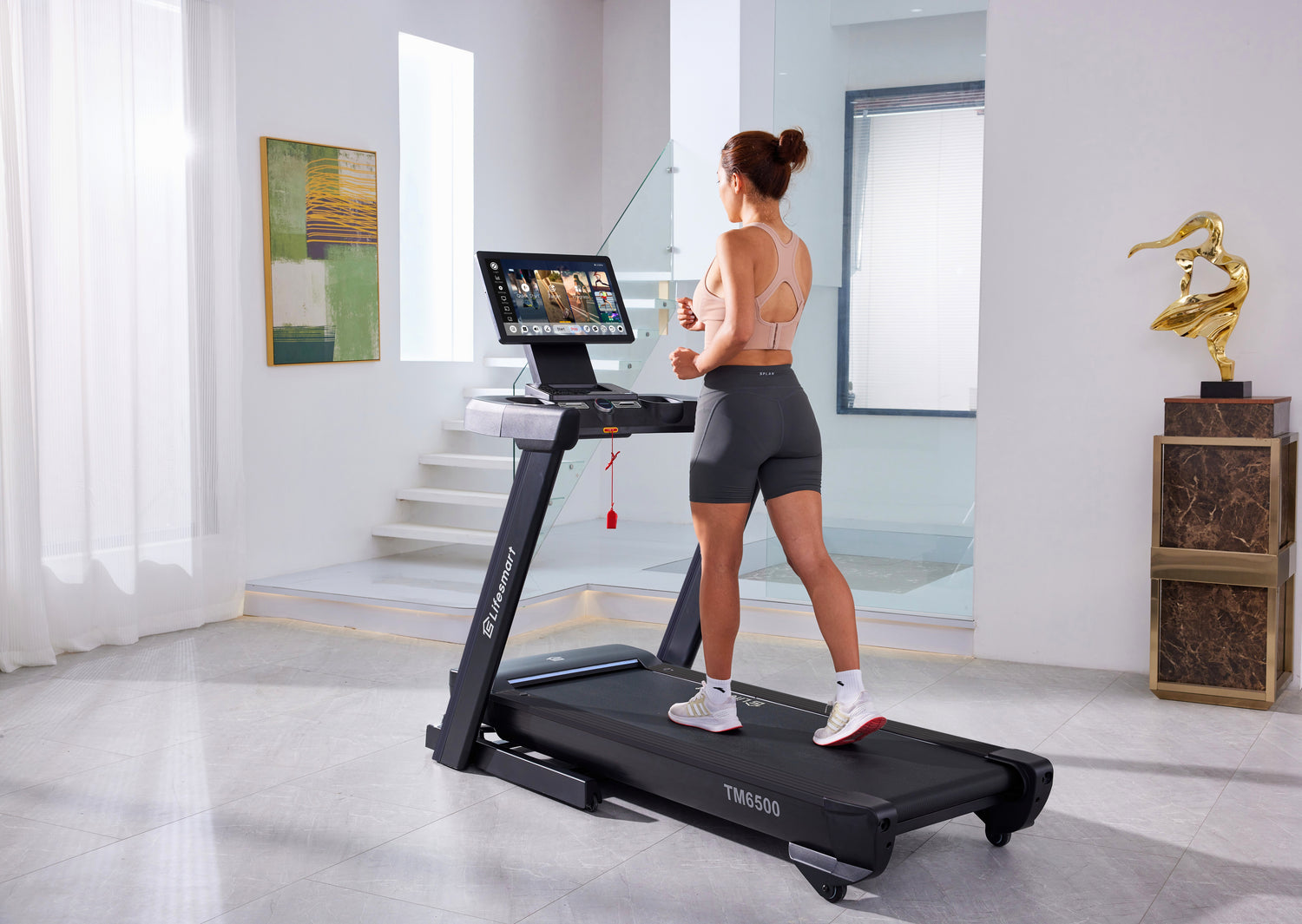 Woman using a Lifesmart TM6500 treadmill in a modern home setting with a laptop on the console.
