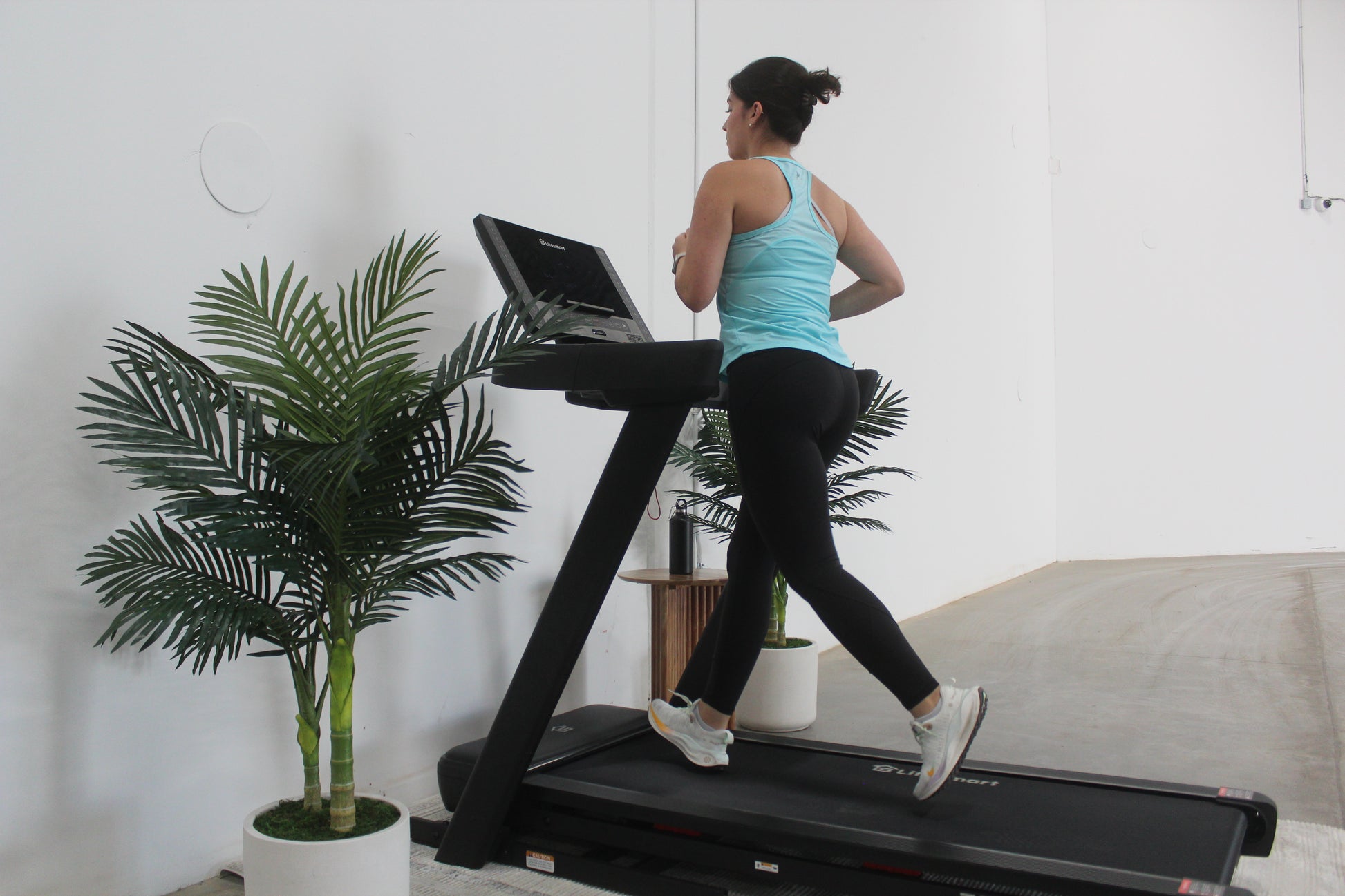 A healthy woman is running on a black Lifesmart TM3000 Power Touch Treadmill indoors, with a plant and a wall in the background.