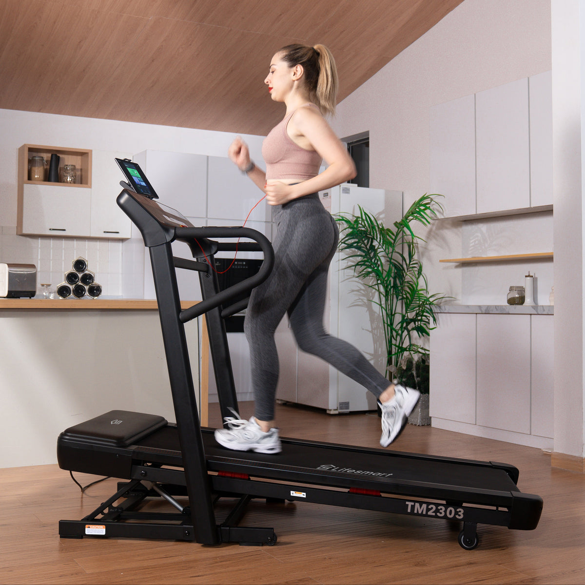 A woman jogging on a black LifeSmarat TM2303 treadmill in a modern home gym with wood floors and a kitchen in the background.