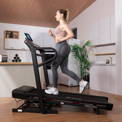 A woman jogging on a black LifeSmarat TM2303 treadmill in a modern home gym with wood floors and a kitchen in the background.