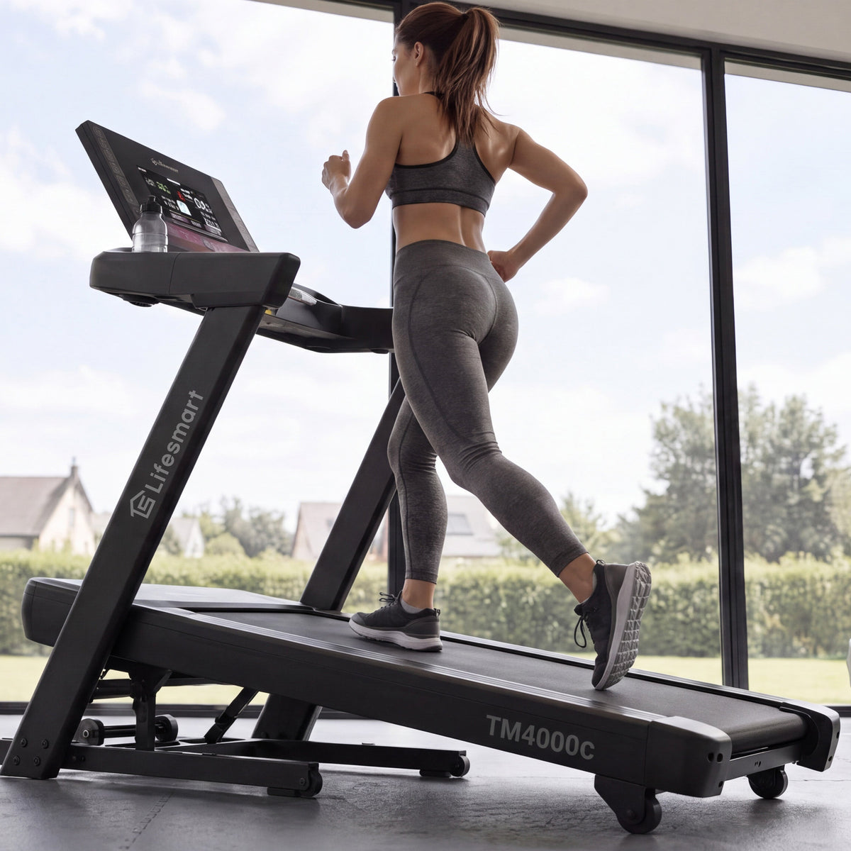 A woman in gray athletic wear runs at incline on a black Lifesmart TM4000C treadmill in a bright gym with large windows and visible dumbbells in the background.